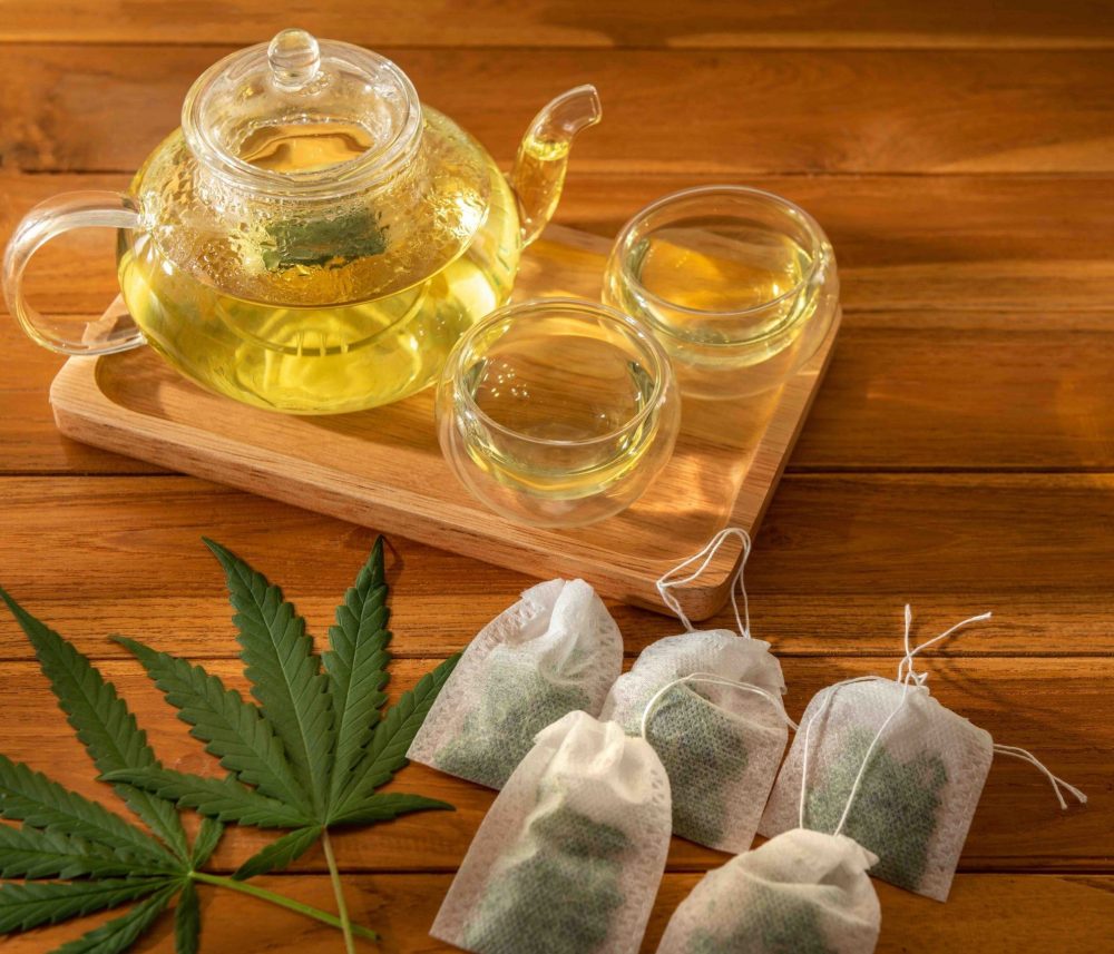 Photo of a pot of hot cannabis tea serves in wooden tray with teabag over wooden table background. Copy space for text banner artwork backdrop design. Eatable recipe health herb vegan drink concept.